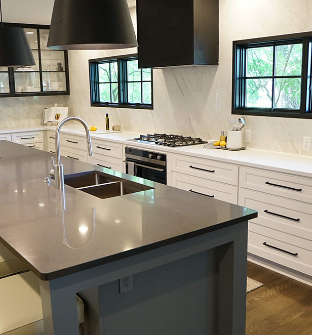 Modern kitchen featuring sleek white cabinets, a gray countertop with a sink, and black pendant lights above the island. Windows reveal greenery outside.