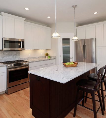 Modern kitchen featuring white cabinetry, stainless steel appliances, a marble countertop island, and wooden floors, with a fruit basket.