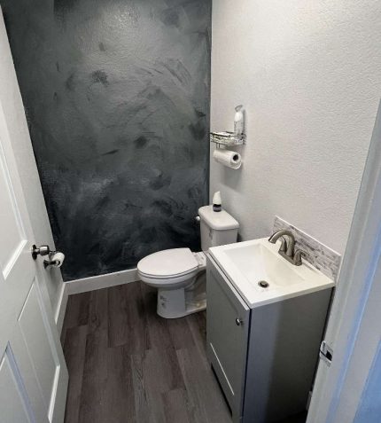 Small modern bathroom featuring a gray accent wall, compact sink cabinet, toilet, and grey wood-like flooring.