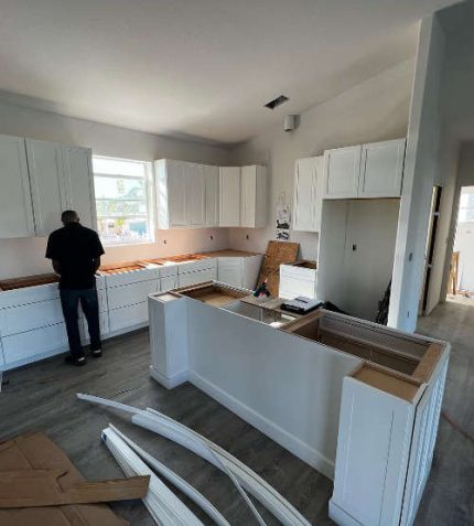 A person assembling white kitchen cabinets in a bright, modern kitchen under construction, surrounded by tools and materials.