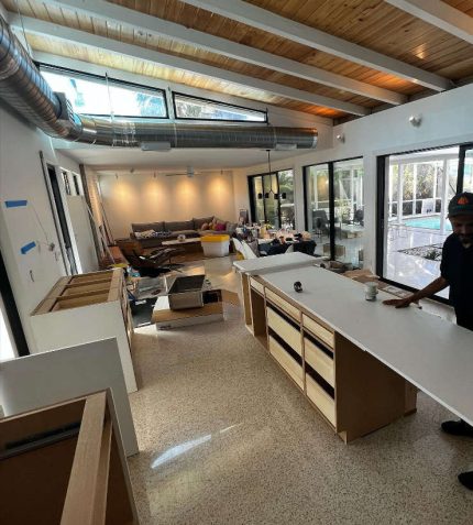 A spacious modern kitchen under construction, featuring a wooden ceiling, large windows, and scattered renovation materials.