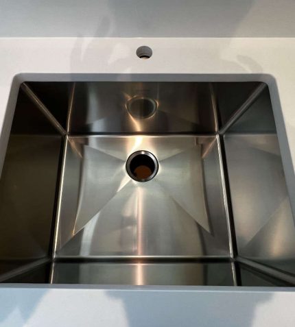 A clean, stainless steel sink with a square design, featuring a central drain hole and smooth, polished surfaces.