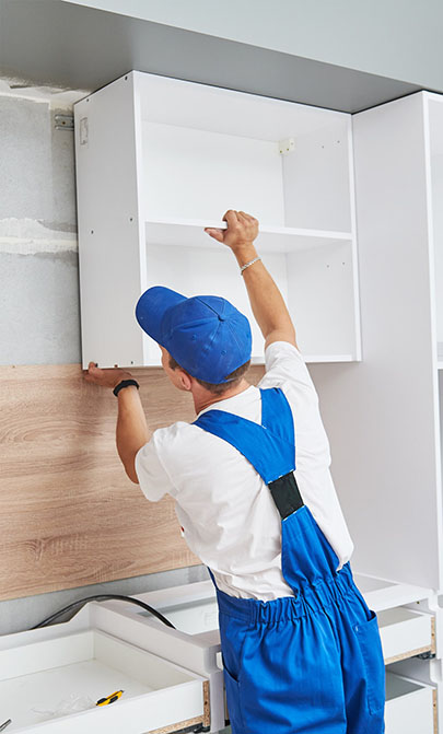 A worker in blue overalls and cap installs a white cabinet in a kitchen, aligning it with the wall and wooden panel below.