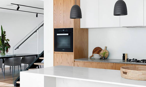 Modern kitchen with wooden accents, a built-in oven, and sleek countertop; features pendant lighting and an open dining area in the background.